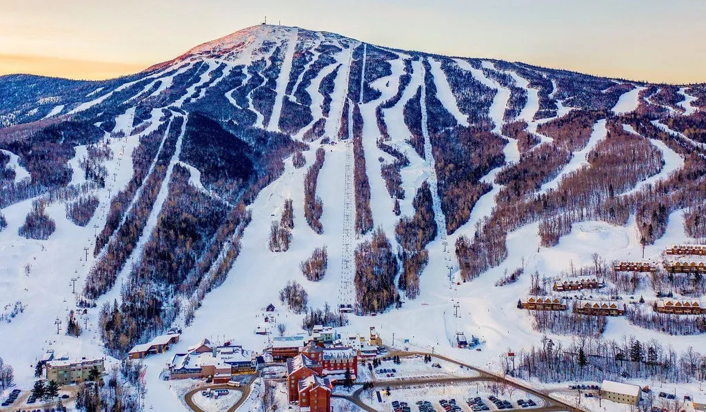 Snowy mountain with ski trails and a village below at sunset.