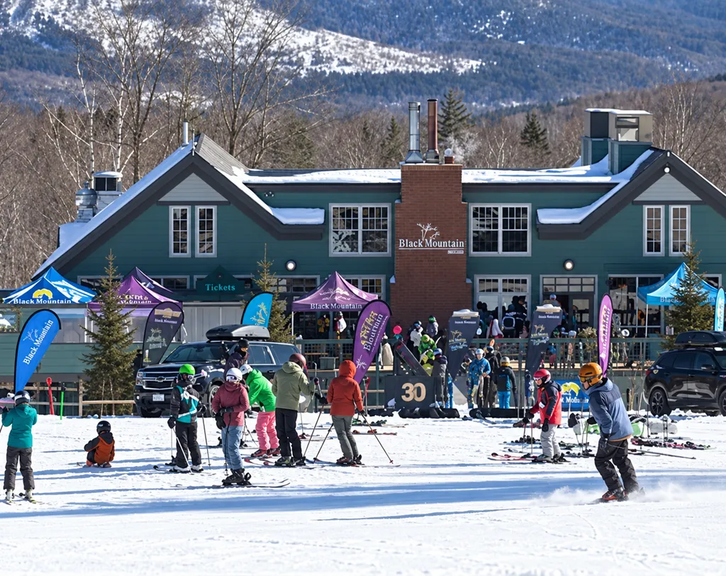 People ski and snowboard in front of a lodge with mountains in the background.